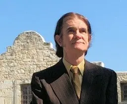 A man with long swept back hair in a brown suit and gold tie standing in front of the Alamo.
