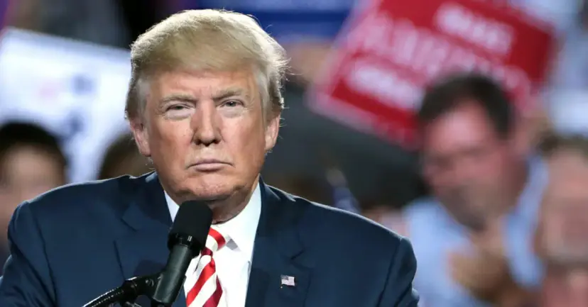 President Donald Trump with a serious expression at a microphone, a crowd holding campaign signs behind him.
