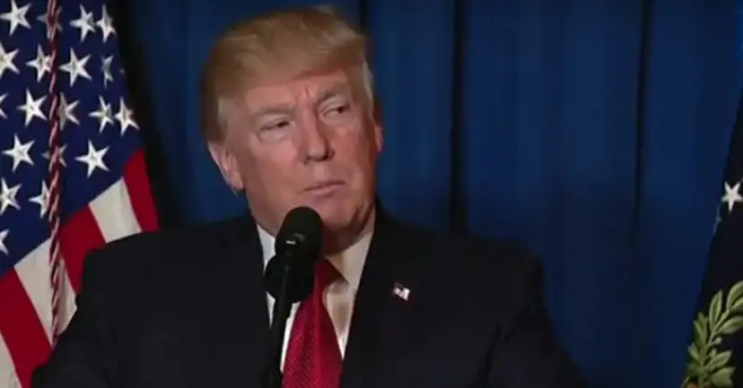 President Donald Trump speaking at a microphone in front of an American flag and a blue curtain.