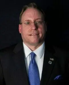 Portrait of a man in glasses, a dark suit and a blue tie against a black background.