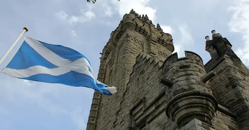 The blue and white Scottish Saltire flies beside a tall stone Gothic tower under a cloudy sky.
