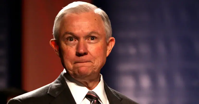 A gray-haired man in a dark suit pressing his lips together against a dark red and blue backdrop.
