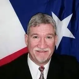 Portrait of a smiling gray haired man with a mustache in front of the Texas flag.