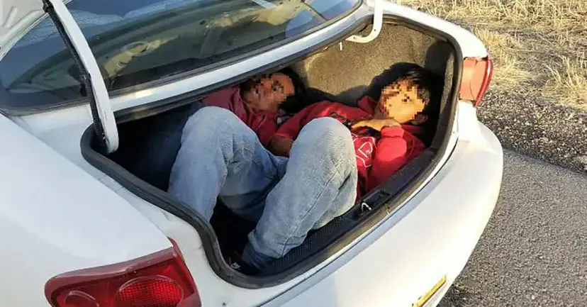 Two people with blurred faces lying hidden in the open trunk of a white car.