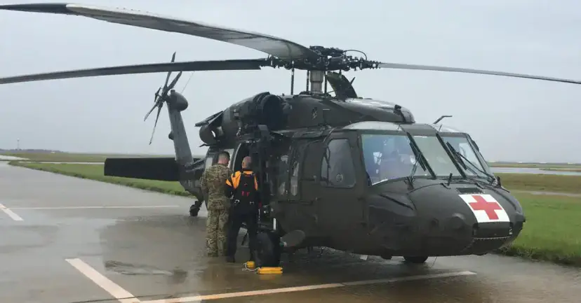 Soldiers boarding a dark green medical helicopter marked with a red cross on a rain-soaked tarmac.