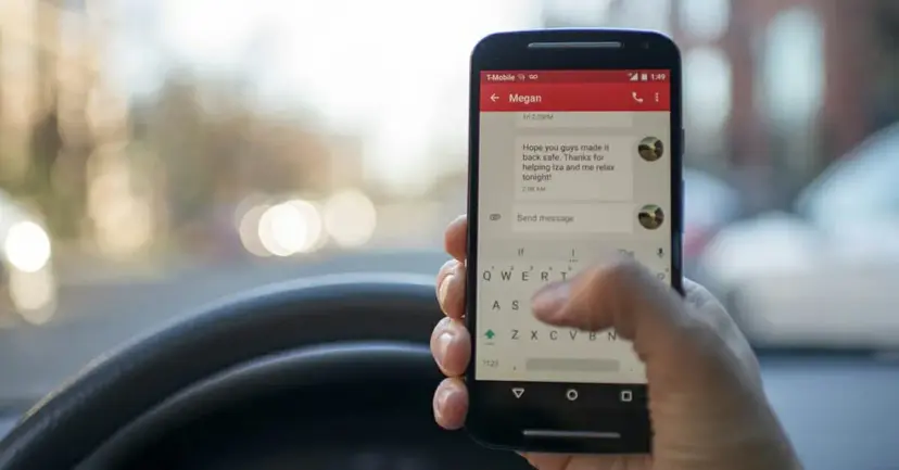 A driver holding a smartphone showing a text conversation above the steering wheel.