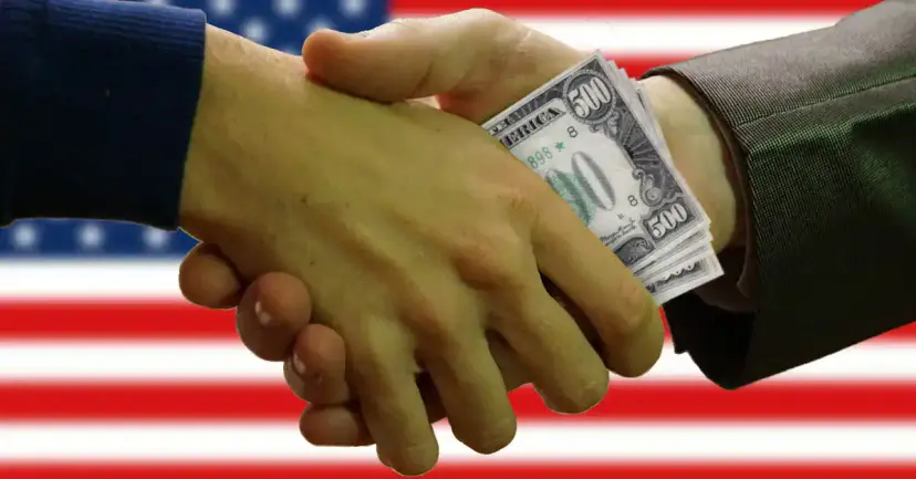 Two hands shaking while passing a stack of 500 dollar bills in front of an American flag.