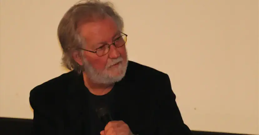 Tobe Hooper, gray-bearded and wearing glasses, holding a microphone at a speaking event.