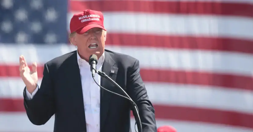 Donald Trump in a red "Make America Great Again" cap speaking at a microphone before an American flag.