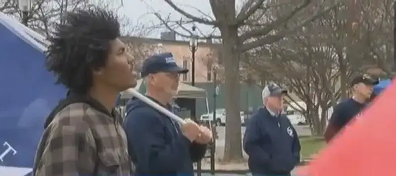 A young man and older men in caps gathered outdoors on a street beside a large flag