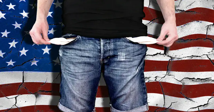 A man in jeans pulling out his empty pockets in front of a cracked American flag.