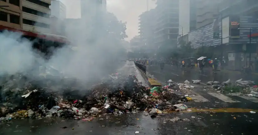 Smoke rising from piles of burning trash on a wet city street lined with high-rise buildings.