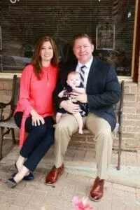 A man in a suit and a woman in a pink top seated on a bench holding a baby.