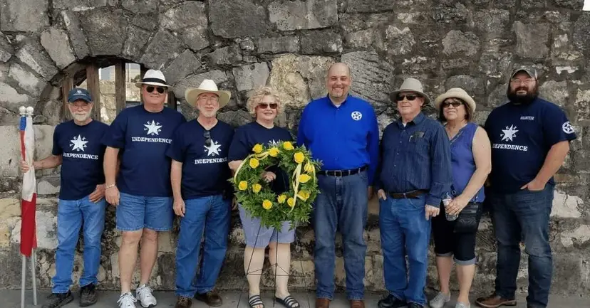 Eight people in Texas independence shirts pose with a yellow flower wreath before a stone wall at the Alamo.