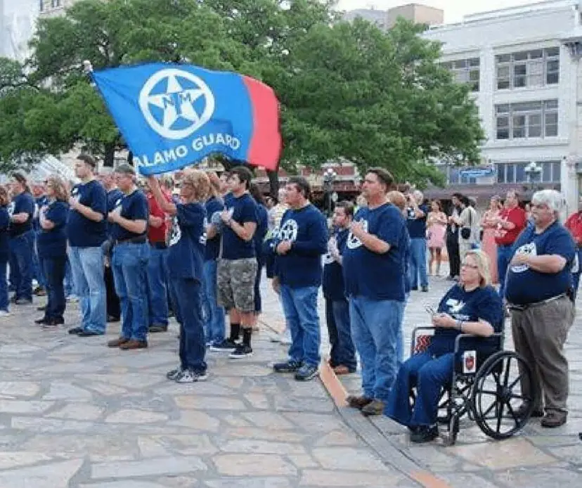 People in navy TNM shirts standing on a stone plaza under a blue "Alamo Guard" flag.