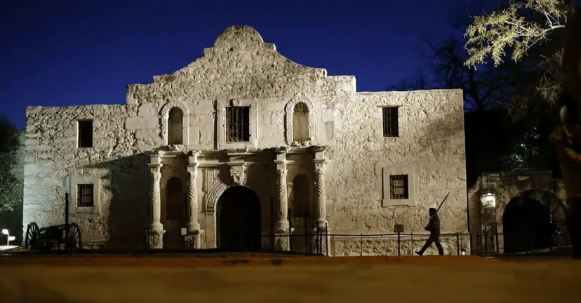 The Alamo chapel lit at night, with a figure carrying a rifle walking past its front.