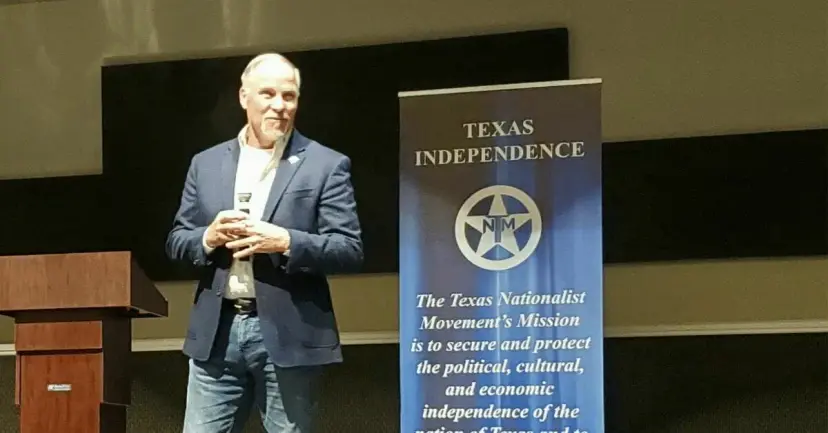 Kyle Biedermann speaking with a microphone on a stage beside a Texas Nationalist Movement banner.