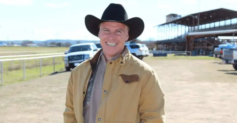 A smiling man in a black cowboy hat and tan jacket standing near a racetrack grandstand.