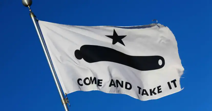 A white 'Come and Take It' flag with a black star and cannon flying against a blue sky.