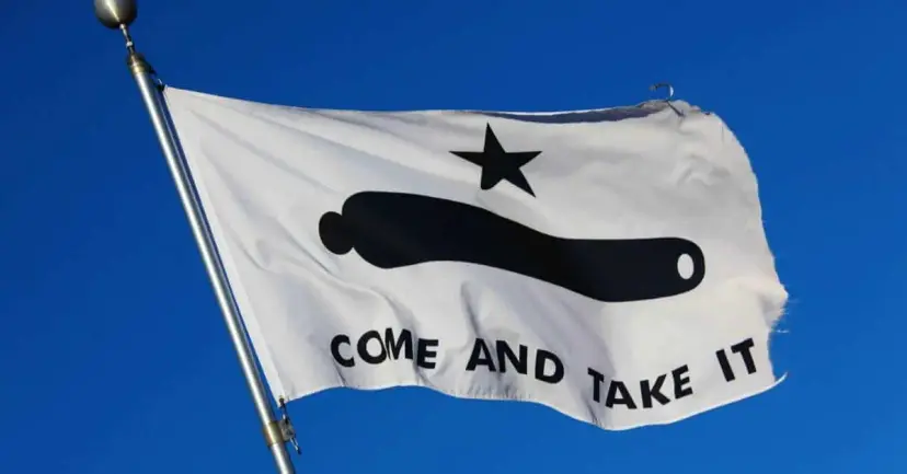 A white 'Come and Take It' flag with a black cannon and star waving against a blue sky.