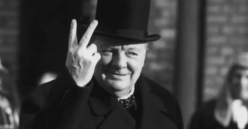 Black-and-white photo of an older man in a top hat and bow tie raising two fingers in a V sign.