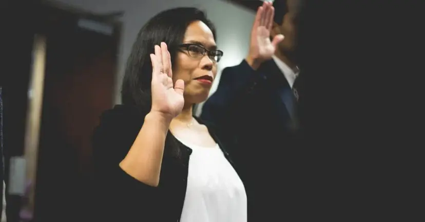 A woman in glasses raises her right hand beside another person as they take an oath.