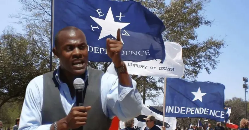 A man speaks into a microphone in front of blue flags reading 'Texas Independence' and 'Liberty or Death.'