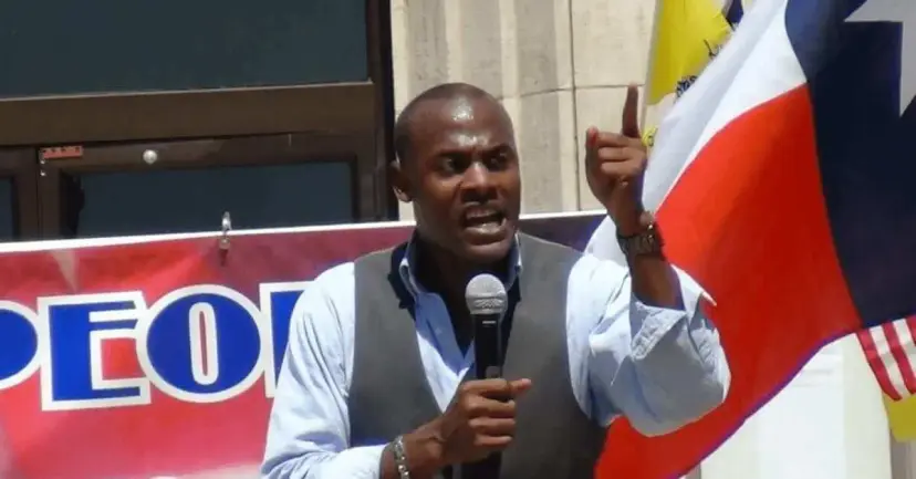 A man in a gray vest speaks into a microphone in front of a Texas flag and a red banner.