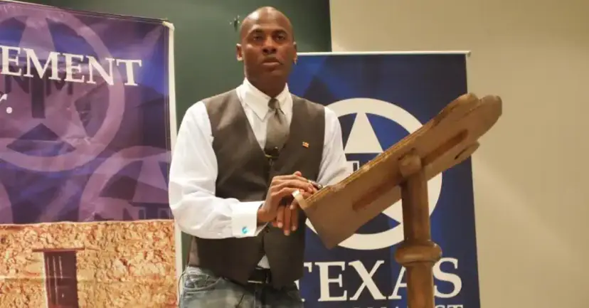 A man in a vest stands at a wooden lectern in front of Texas Nationalist Movement banners.