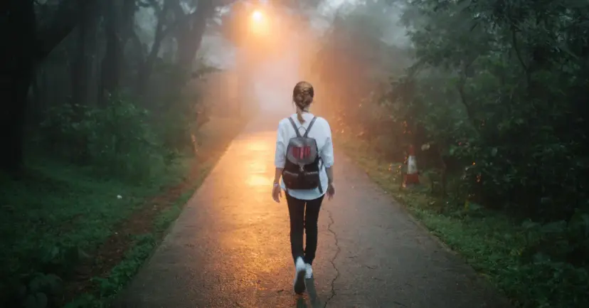 A woman with a backpack walking down a misty forest path toward a glowing light.
