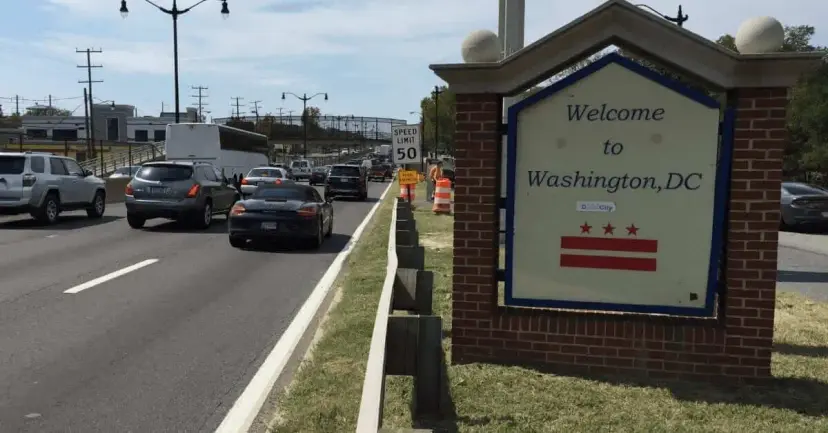 A 'Welcome to Washington, DC' sign beside a busy multi-lane road lined with traffic.