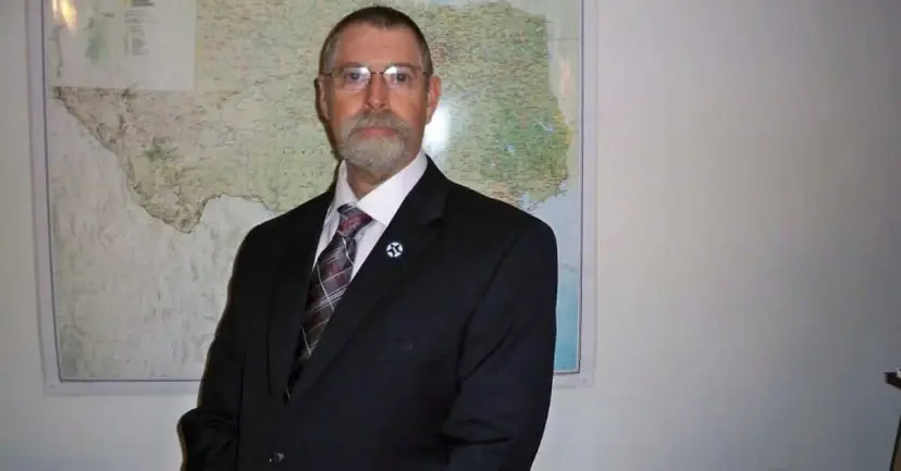 A bearded man in glasses and a dark suit standing in front of a large wall map of Texas.