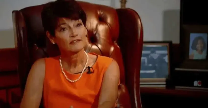 Woman with short dark hair in an orange dress and pearls, seated in a leather office chair during an interview.