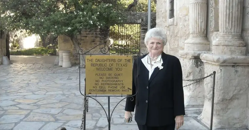Older woman in a dark suit standing beside a Daughters of the Republic of Texas welcome sign at the Alamo.