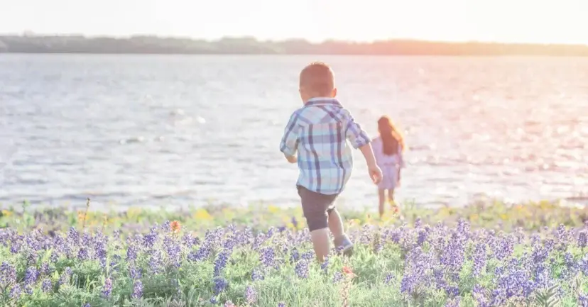 Two children walk through a field of bluebonnets toward a lake.