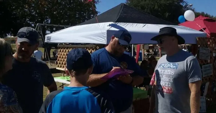 Volunteers talk with visitors and fill out a clipboard at an outdoor festival booth under a canopy.
