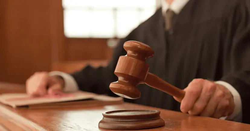 A judge in a black robe raising a wooden gavel over its sound block at a courtroom bench.
