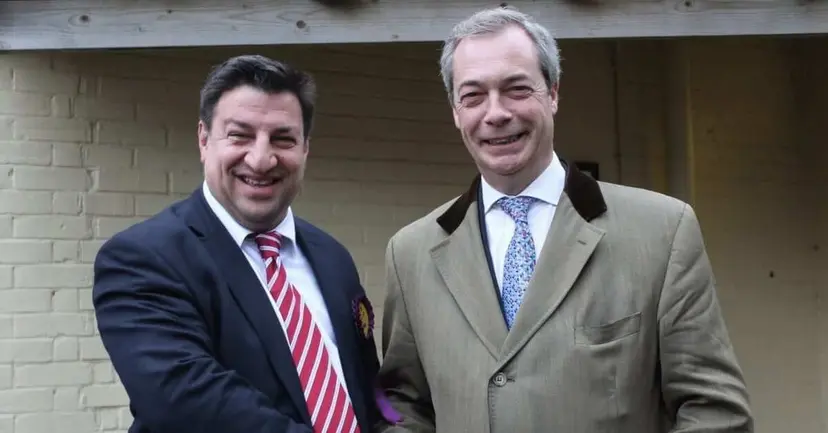 Two smiling men in suits shaking hands in front of a white brick wall.