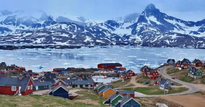 Brightly painted houses of a coastal Greenland town above an icy bay, with snow-capped mountains behind.