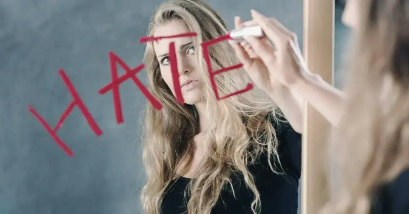 A woman looks at a mirror as a hand writes the word 'hate' in red across the glass.