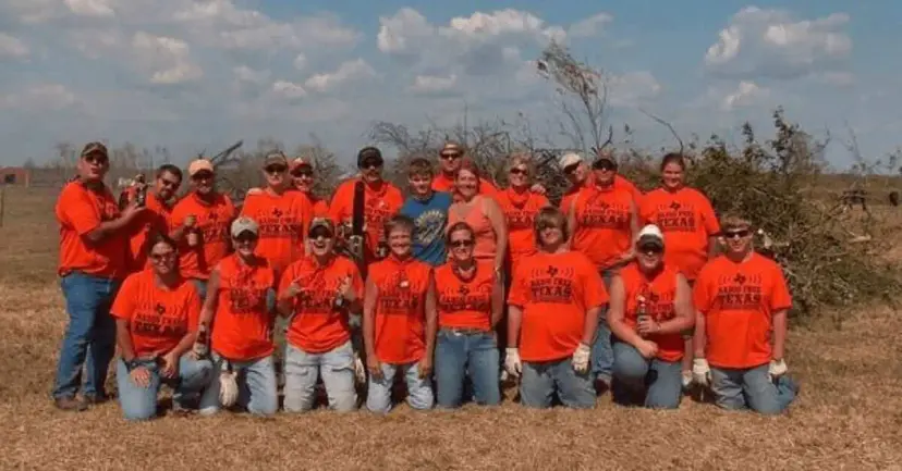 A group of volunteers in matching orange Texas shirts posing in a field among storm debris and broken trees.