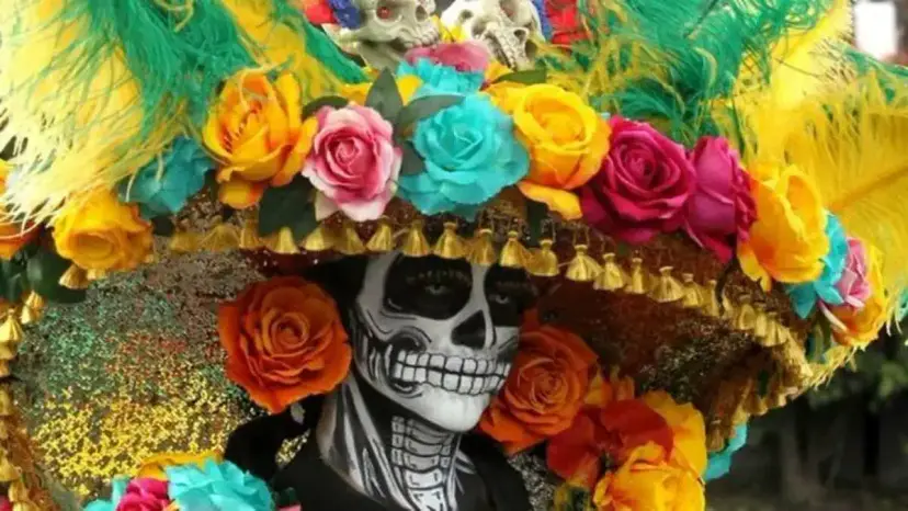 A person in skull face paint wearing a large hat covered in bright flowers and feathers.