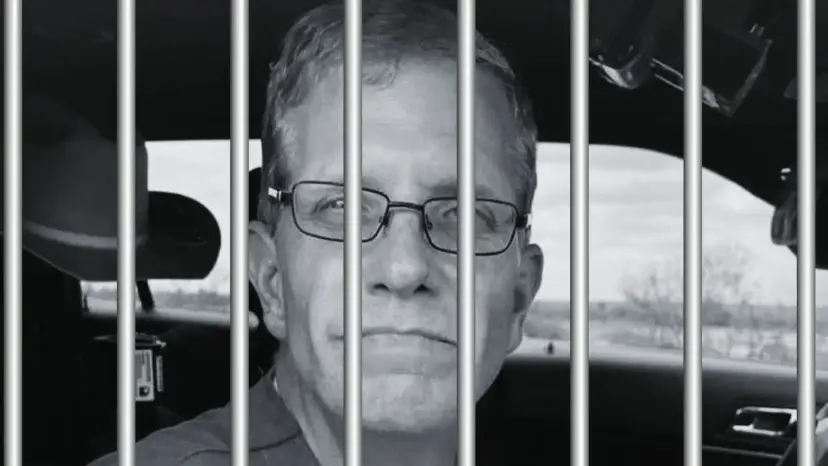 Black and white photo of a man with glasses behind jail bars, seated in a patrol car.