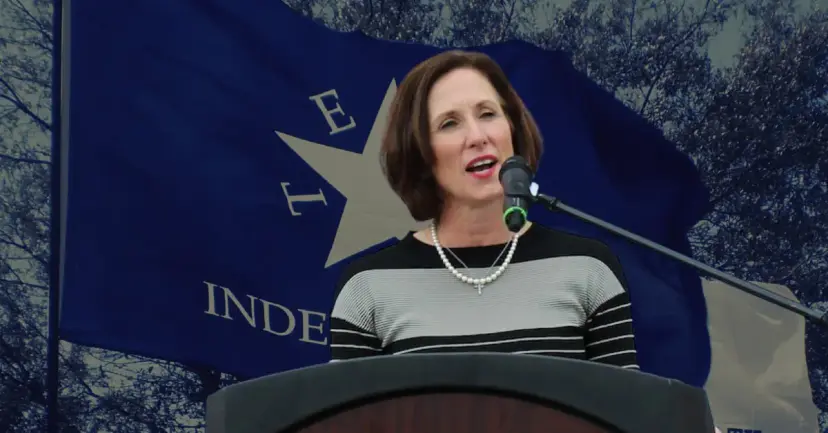 Senator Lois Kolkhorst speaks at a podium microphone in front of a blue Texas Independence flag.