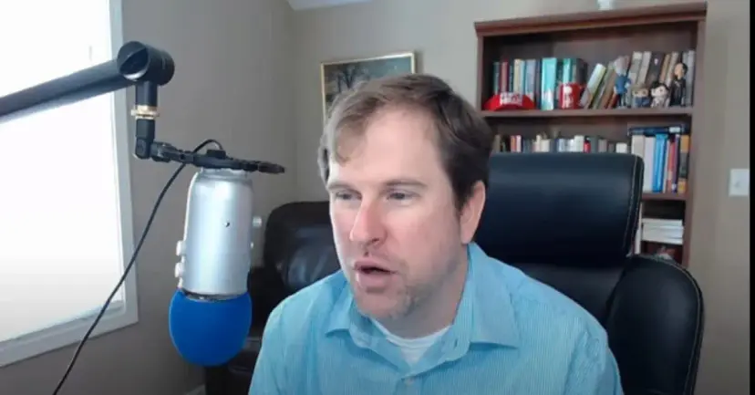A man in a blue shirt speaks into a podcast microphone in a home office with bookshelves.