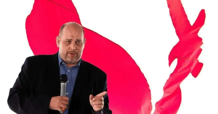 Daniel Miller speaking into a handheld microphone in front of a large red flag.