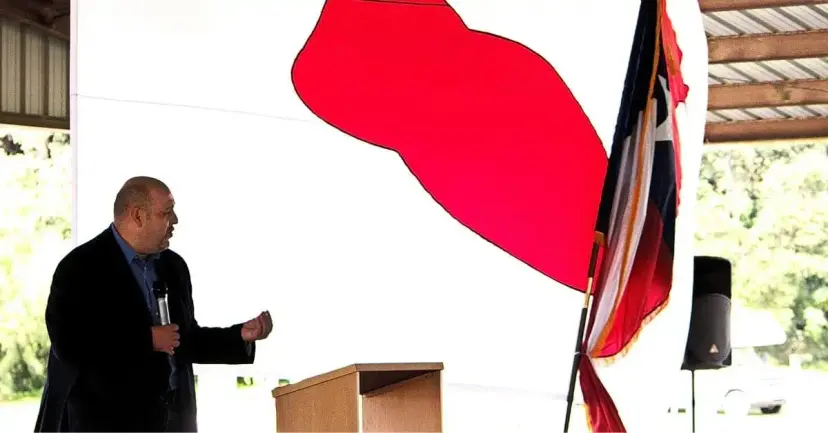 Daniel Miller speaks with a microphone near a podium and a Texas flag in an open pavilion.