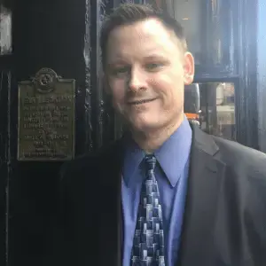 Nate Smith in a dark suit and blue shirt standing beside a historical plaque on a dark doorway.