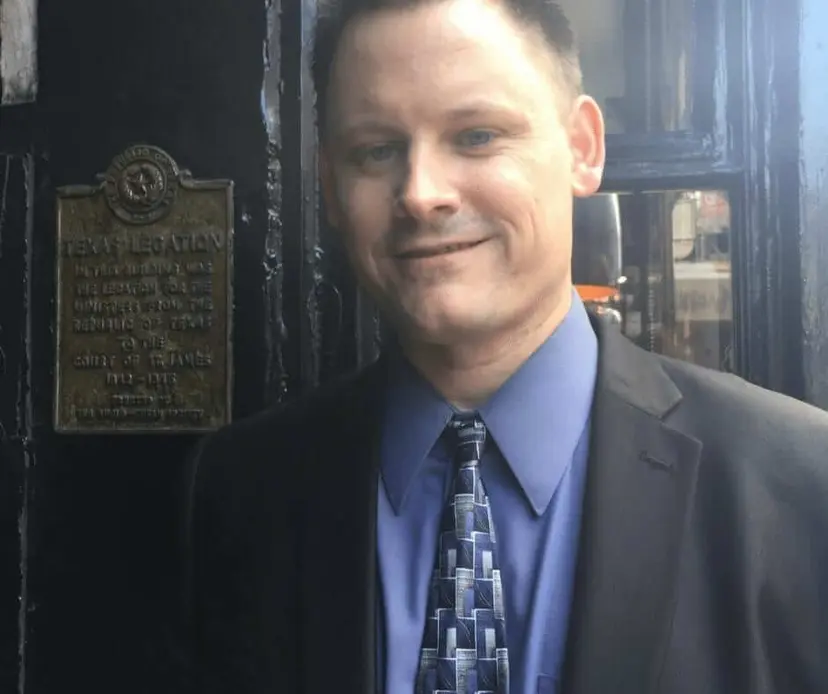 Smiling man in a suit and blue tie standing beside a bronze plaque marking a former Texas legation.