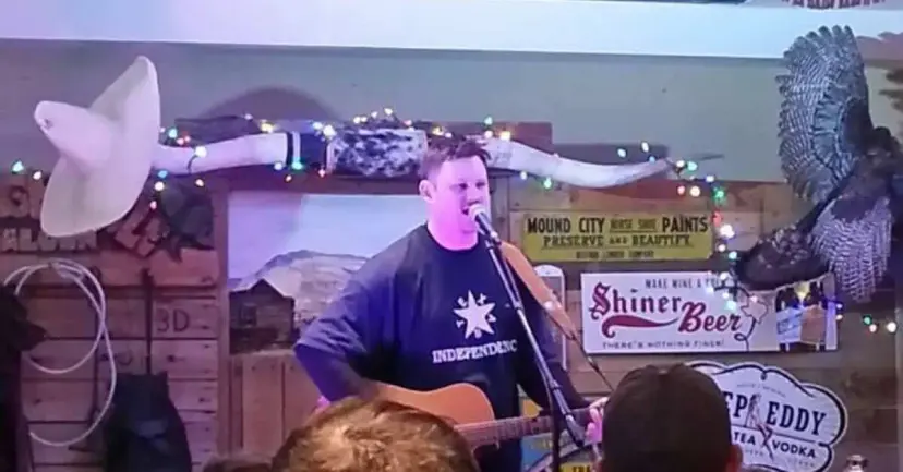 Man in an independence T-shirt singing and playing acoustic guitar on a small stage decorated with longhorns and lights.